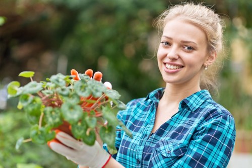 A young woman with blonde hair tied back, smiling and wearing a blue plaid shirt and white gardening gloves, is tending to a potted plant with green and reddish leaves, set outdoors in a garden area. The background features lush greenery with blurred foliage, indicating a well-maintained garden space in Crouch End. The scene captures a moment of garden maintenance or planting activity, reflecting professional gardening practices suitable for a residential outdoor environment within the vicinity of N8 postcode area. The natural daylight highlights the vibrant plant and the woman's cheerful expression, aligning with gardening and landscaping services offered by Garden Maintenance Crouch End in North London. This detailed, naturalistic image emphasizes garden care, plant handling, and outdoor upkeep typical of local garden maintenance work in the Crouch End district.